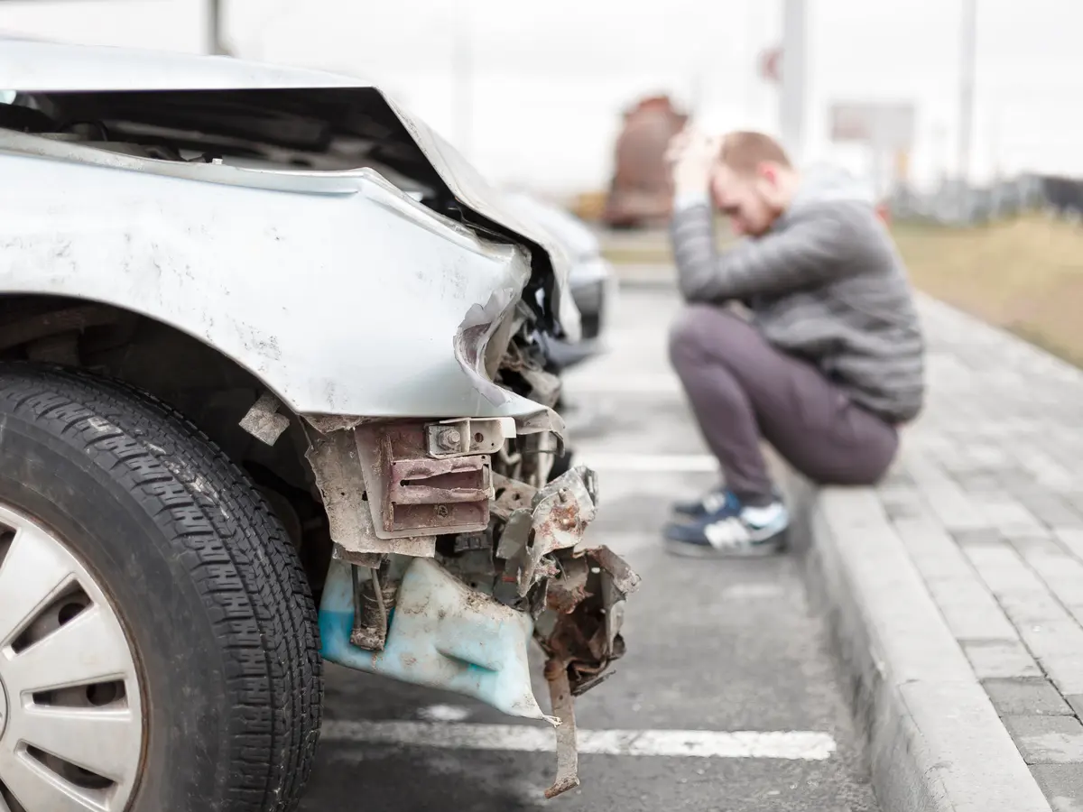 Driver sitting on a curb holding his head next to a car damaged in a collision